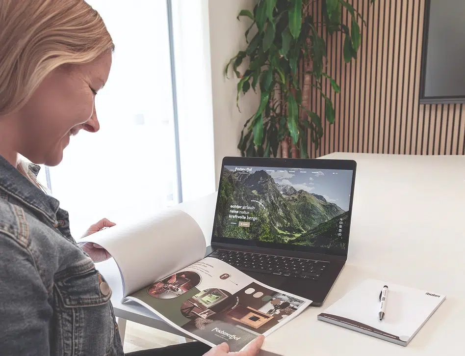 Eine Frau lächelt über eine Zeitschrift und einen Laptop mit Hotel-Branding-Design auf einer Berg-Website.